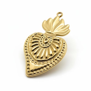 Op de afbeelding: Een goudkleurige hanger in de vorm van een hart, met ingewikkelde reliëfdetails. De hanger heeft een gestileerd vlamontwerp aan de bovenkant en een lus om op te hangen. Het hart is versierd met een spiraal- en stippenpatroon.