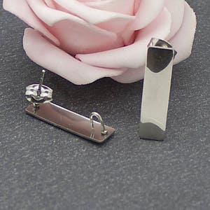 May include: A pair of contemporary silver earrings. Each earring features a rectangular design with a dark grey accent at the base and a silver post. The earrings are presented on a dark grey surface with a pink rose in the background.