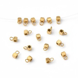 Peut inclure: Des apprêts de bijoux en métal doré, comprenant de petits cubes avec des boucles attachées. Les apprêts sont dispersés sur une surface blanche. Ces composants sont utilisés dans la fabrication de bijoux.