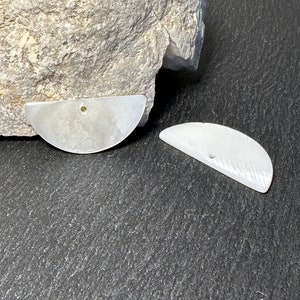 Peut inclure: Deux pendentifs nacrés en forme de demi-lune avec un petit trou percé près du sommet. Les pendentifs ont un éclat subtil et irisé et sont présentés sur un fond texturé gris avec une grosse pierre.