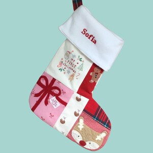 May include: A patchwork Christmas stocking featuring a white cuff embroidered with the name Sofia in red. The body displays a My First Christmas graphic, a pink section with a red bow, a white strip with small puddings, and a reindeer face.