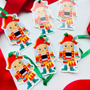 May include: Six colorful nutcracker ornaments with red hats, white faces, and black eyes. Each ornament has a red and gold drum and is holding a drumstick. The ornaments are on a white background.