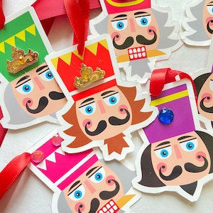 Puede incluir: Seis adornos de cascanueces coloridos con coronas doradas y ojos azules. Los adornos están hechos de papel y tienen un tema festivo navideño.