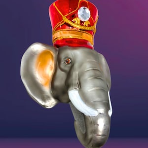 May include: A decorative elephant head ornament with a red and gold hat, a large crystal, and a gold ribbon for hanging. The elephant is gray with white tusks and a detailed trunk. The ornament is approximately 4 inches tall.