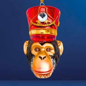 Könnte beinhalten: Dekorative Weihnachtsbaum-Ornament in Form eines Affenkopfes. Der Affe trägt einen roten Hut mit goldenen Details und einem klaren Edelstein. Das Gesicht des Affen ist gold und braun, mit einem lächelnden Ausdruck. Ein goldenes Band ist befestigt.