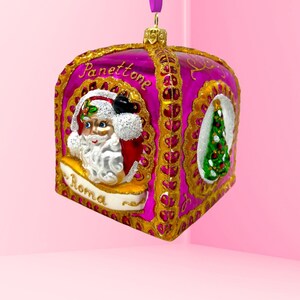 Puede incluir: Un adorno navideño de vidrio rosa y dorado con forma de caja con un diseño de Papá Noel y un árbol de Navidad. El adorno tiene las palabras "Panettone" y "Roma" en él.