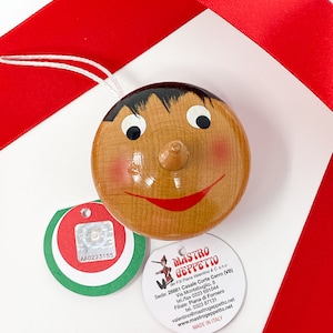 May include: A wooden toy head with a painted face, brown hair, and a red smile. The toy is attached to a string and has a green and red tag with the text "AA0223155". A white tag with the text "MASTRO GEPPETTO" is also attached to the toy.