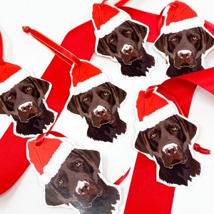 May include: Five Christmas ornaments featuring brown Labrador Retriever dogs wearing Santa hats. The ornaments are white with red and white Santa hats.
