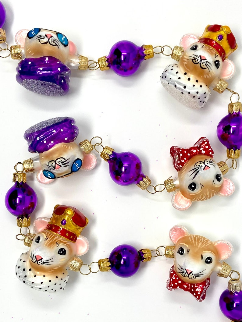K&ouml;nnte beinhalten: Eine violette und goldene Weihnachtsgirlande mit Glasornamenten, die M&auml;use zeigen, die verschiedene H&uuml;te und Accessoires tragen. Die Ornamente sind mit einer goldenen Kette aneinandergereiht.