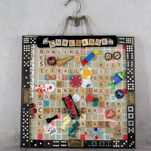 May include: A framed game-themed collage featuring Scrabble tiles spelling "CHALLENGE", "BOWLING", "CHESS", and "STARS". The collage includes game pieces like poker chips, dominoes, and a toy car. A metal hanger is attached at the top.