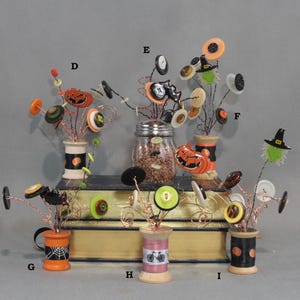 May include: A collection of decorative wire sculptures featuring colorful buttons and Halloween-themed charms, each set in a wooden spool base. The arrangements include pumpkins, bats, and witch hats, with letters D, E, F, G, H, and I labeling each piece.