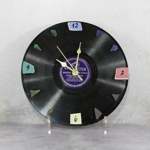 May include: A black vinyl record clock with colorful, geometric hour markers. The clock features gold-colored hands and a purple and white label in the center. The clock is displayed on a clear stand.