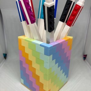 Puede incluir: Un portalápices colorido hecho de bloques entrelazados, lleno de bolígrafos. El portalápices presenta un patrón de chevrón arcoíris. Los bolígrafos tienen barriles blancos y detalles coloridos en rojo, negro, azul, morado y turquesa.