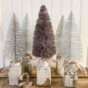 May include: A collection of decorative miniature Christmas trees. The trees are in shades of gray and brown, with a distressed white base. A silver bell with a pink ribbon is also present.