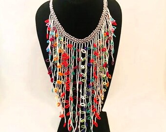May include: A long, colorful beaded necklace with a cascading design. The necklace is made of white beads with small, colorful beads interspersed throughout. The necklace is displayed on a black mannequin.