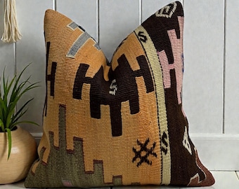 May include: A decorative pillow with a geometric pattern in shades of brown, orange, and pink. The pillow has a textured appearance and is likely made of woven fabric. The design features repeating shapes and symbols, creating a vintage or bohemian aesthetic.