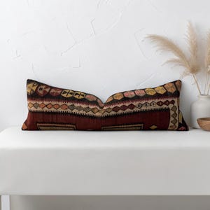 May include: A rectangular lumbar pillow featuring a woven textile cover with a deep red base color. The fabric displays a repeating geometric pattern in shades of yellow, black, and tan, including diamond shapes and circular motifs along the top edge.