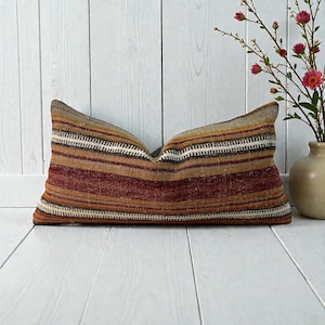 May include: A rectangular cushion with a striped design in red, brown, and gold hues. The cushion has a textured look and is set on a white wooden surface. A vase with pink flowers is in the background.