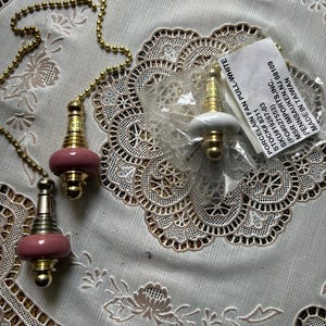 May include: A set of porcelain fan pull chains with a gold finish and pink accents. One pull is still in its original packaging. The pulls are on a white lace tablecloth.