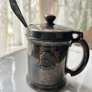 May include: Silver sugar dispenser with a hinged lid and a small spoon. The dispenser has a crest with a crown on it.