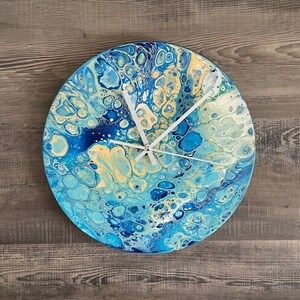 May include: A round clock with a blue, yellow, and white abstract paint design. The clock hands are white.