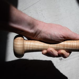 Op de afbeelding: Een houten honkbalknuppel met een lichtbruine afwerking en een afgeronde handgreep. De knuppel wordt in een hand gehouden, waarbij de natuurlijke houtnerf te zien is. De knuppel lijkt nieuw en ongebruikt.