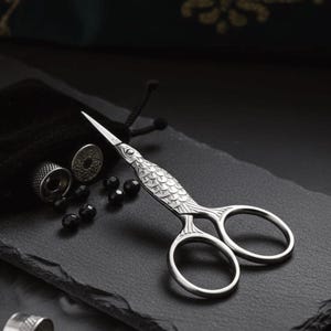 Może przedstawiać: Srebrne nożyczki do haftu z motywem w kształcie ryby, ze szczegółowym wzorem łusek na uchwycie. Nożyczki są prezentowane na ciemnej, teksturowanej powierzchni, obok małego czarnego woreczka, naparstków i czarnych koralików.