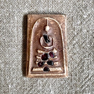 May include: A brown ceramic amulet with a white design and red glass accents. The amulet is shaped like a small rectangular box with a rounded top.