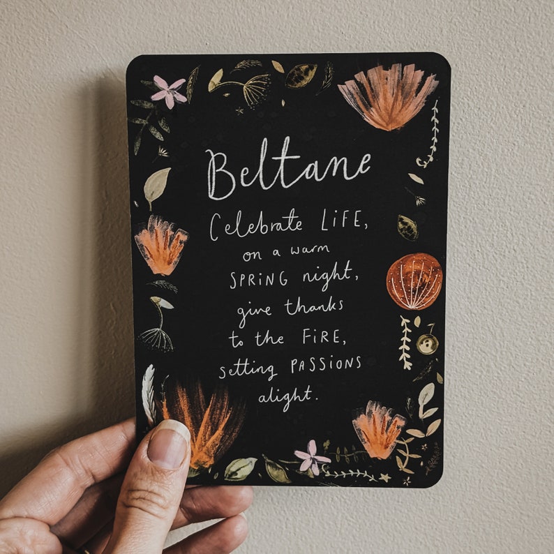 Pu&ograve; includere: Carta nera con testo bianco e illustrazioni floreali. Il testo recita "Beltane Celebra la VITA, in una calda notte di PRIMAVERA, ringrazia il FUOCO, accendendo le PASSIONI."