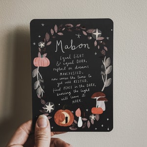 May include: A black card with a white illustration of a pumpkin, leaves, and stars. The text on the card reads "Mabon Equal LIGHT & equal DARK, reflect on dreams MANIFESTED, now comes the time to get well RESTED, find PEACE in the DARK, knowing the light will leave its MARK."