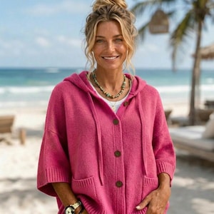 May include: A bright pink knit cardigan featuring a hood, short sleeves, and a front button closure with four visible buttons. The garment includes two front patch pockets and is worn over a plain white shirt, paired with a beaded necklace.