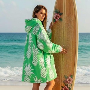 Pode incluir: Um poncho de praia verde com capuz e um padrão de abacaxi branco. O poncho é usado numa praia com uma prancha de surf. A prancha de surf é decorada com desenhos florais. O oceano está ao fundo.