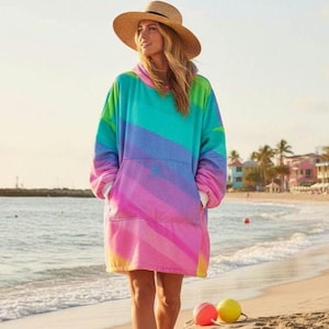 Pode incluir: Um cobertor com capuz oversized em tons de arco-íris de azul, verde, rosa e roxo, com um bolso canguru. O cobertor é usado na praia. Um chapéu de palha também é visível.