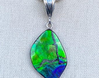May include: A pendant necklace featuring a vibrant, iridescent gemstone in shades of green, blue, and purple. The gemstone is set in a silver-colored metal setting and hangs from a silver chain. The gemstone has a unique, organic shape.