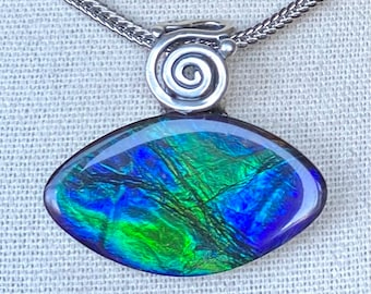 Può includere: Collana con pendente con una gemma iridescente nei toni del blu e del verde. La gemma è incastonata in metallo argentato con un motivo a spirale, collegata a una catena d'argento. Il pendente ha una forma ovale.