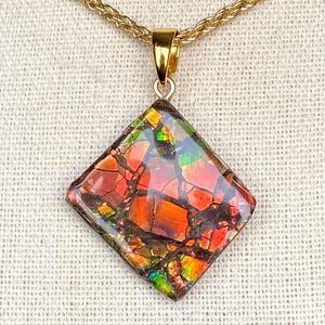 May include: A diamond-shaped pendant with a vibrant, multicolored gemstone, featuring shades of red, orange, green, and brown. The pendant is suspended from a gold-colored chain. The gemstone has a cracked, mosaic-like appearance.