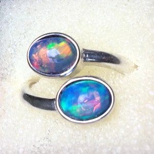 May include: Two silver rings, each featuring an oval-shaped opal gemstone. The opals display a vibrant play of color, with hues of blue, green, orange, and red. The rings are set against a white background.