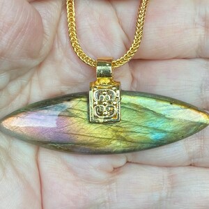 May include: A gold chain necklace with a labradorite pendant. The pendant is a long, oval-shaped stone with iridescent green, blue, and gold hues. The pendant is attached to the chain with a gold bail that has a decorative design.