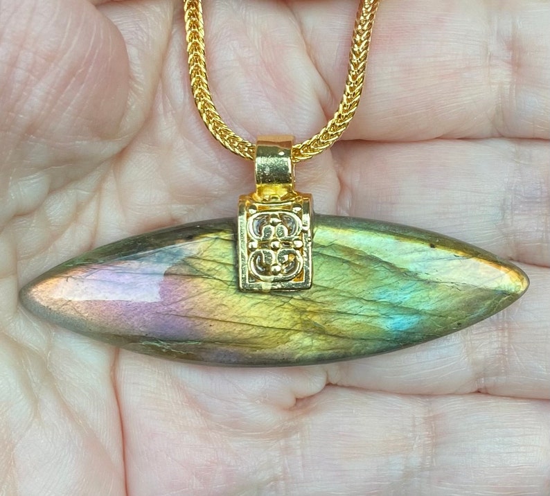 May include: A gold chain necklace with a labradorite pendant. The pendant is a long, oval-shaped stone with iridescent green, blue, and gold hues. The pendant is attached to the chain with a gold bail that has a decorative design.