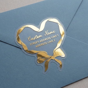 May include: A clear sticker with gold foil detailing, shaped like a heart with a ribbon and bow design. The sticker has the text "Custom Name", "Street Address Line 1", and "Address Line 2". It is placed on a blue envelope.