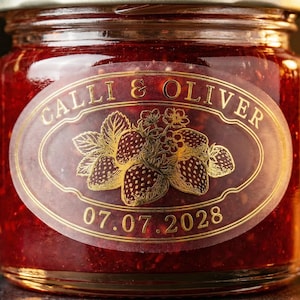 Può includere: Un barattolo di marmellata rossa con un'etichetta dorata con la scritta "Calli & Oliver" e un'illustrazione di fragole. La data "07.07.2028" è stampata sotto. Il barattolo ha un coperchio argentato.