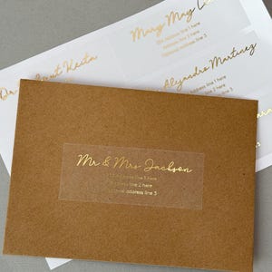 Peut inclure: Un autocollant rectangulaire transparent avec une inscription dorée "Mr & Mrs Jackson" et un espace pour une adresse. L'autocollant est sur une enveloppe brune.