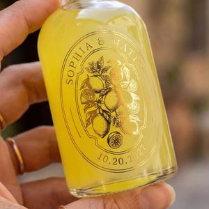 May include: A clear glass bottle filled with yellow liquid, with a gold label displaying the names "SOPHIA & MATISS" and the date "10.20.2027". The label features a gold illustration of lemons and leaves. The bottle is held in a hand.