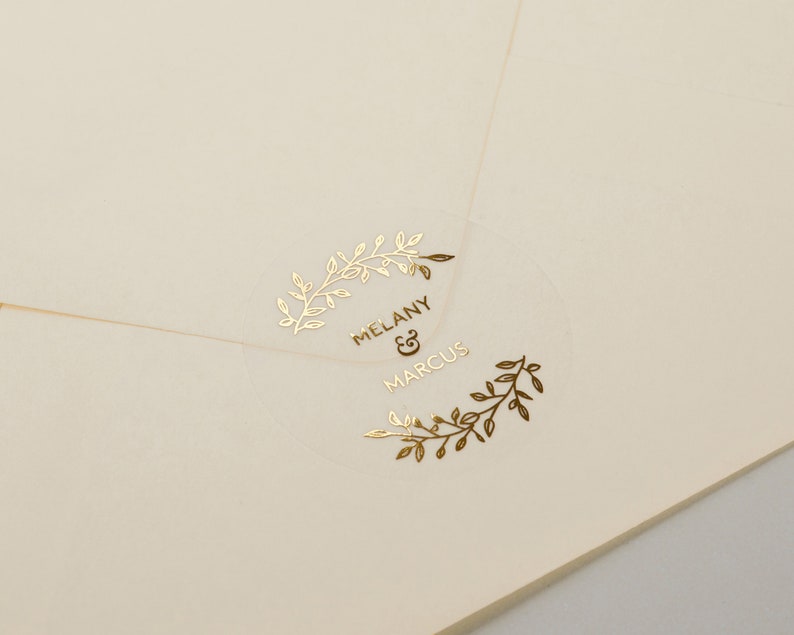 以下が含まれることがあります： 葉のリースと「Melany & Marcus」の文字が書かれた金箔のデザインが施された透明なステッカーです。