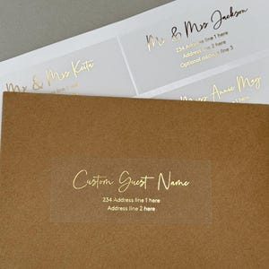 Op de afbeelding: Transparante, rechthoekige, zelfklevende etiketten met gouden folielettering. De etiketten zijn ontworpen voor het adresseren van bruiloften of evenementen. De tekst op de etiketten luidt "Mr. & Mrs. Keita", "Mr. & Mrs. Jackson", "Mrs. Annie May" en "Custom Guest Name".