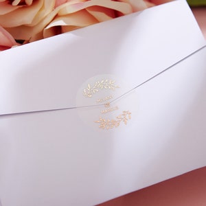 以下が含まれることがあります： ゴールドの花柄と「Melany & Marcus」の文字が書かれた透明なステッカーが貼られた白い封筒です。