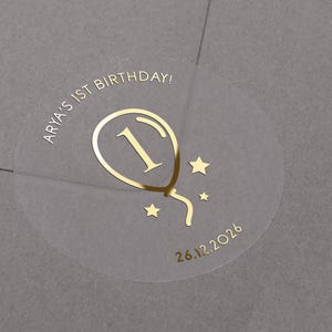 May include: A clear round sticker with gold foil lettering and a balloon graphic. The sticker says "Arya's 1st Birthday!" and "26.12.2026".