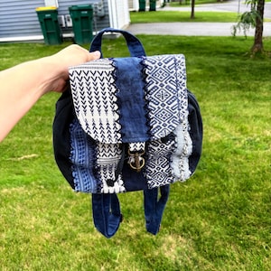 May include: A small backpack featuring a patchwork design of dark blue and white fabric with various geometric patterns. The bag includes a top handle, adjustable shoulder straps, and a front flap secured with a metal lobster clasp closure.