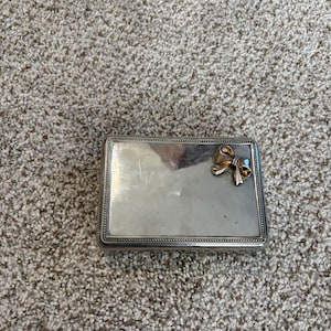May include: A rectangular silver-toned metal box with a polished, reflective surface and a beaded border. A small, three-dimensional gold-toned metal bow ornament is attached to the upper right corner of the lid, adding a decorative accent.