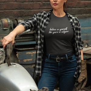 May include: A woman wearing a black t-shirt with the text "believe in yourself like you're a religion". She is wearing blue jeans, a black and white plaid shirt, and a welding helmet. She is leaning on a motorcycle.